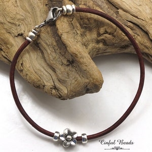 May include: A brown leather bracelet with a silver flower charm and silver beads. The bracelet is on a wooden background. Cinfula Beads.