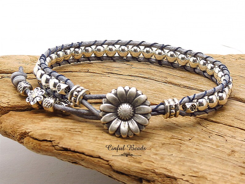 K&ouml;nnte beinhalten: Ein graues Lederarmband mit silbernen Perlen und einem silbernen G&auml;nsebl&uuml;mchen-Anh&auml;nger. Das Armband ist doppelschichtig und hat einen blauen Faden, der durchgewebt ist. Das Armband liegt auf einer Holzoberfl&auml;che.