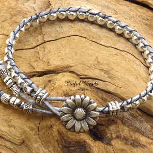 K&ouml;nnte beinhalten: Ein graues Lederarmband mit silbernen Perlen und einem silbernen G&auml;nsebl&uuml;mchen-Anh&auml;nger. Das Armband ist mit einem Knoten gebunden und hat einen blauen Faden, der durchgeflochten ist. "Cinfu Beads" steht auf der Holzoberfl&auml;che.