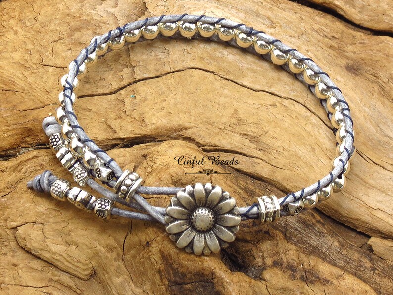 K&ouml;nnte beinhalten: Ein graues Lederarmband mit silbernen Perlen und einem silbernen G&auml;nsebl&uuml;mchen-Anh&auml;nger. Das Armband ist mit einem Knoten gebunden und hat einen blauen Faden, der durchgeflochten ist. "Cinfu Beads" steht auf der Holzoberfl&auml;che.