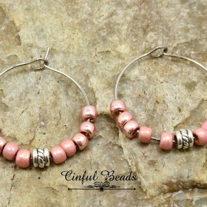 May include: A pair of silver hoop earrings with pink beads and a silver bead accent. The earrings are on a textured brown surface. "Cinfu Beads" is written on the surface.