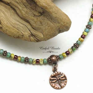 May include: A beaded necklace with a copper dragonfly pendant. The necklace features a mix of colorful beads in shades of green, yellow, and burgundy. The pendant is round with a dragonfly design.