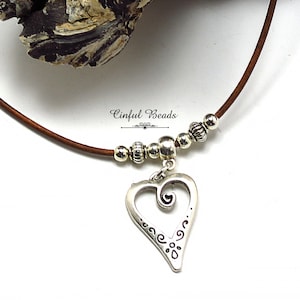 May include: A brown leather cord necklace with silver beads and a silver heart pendant. The heart pendant has a swirl design and is engraved with a floral pattern.