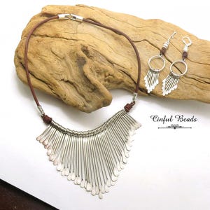 May include: A silver-toned necklace and matching earrings set. The necklace features a brown leather cord and a silver fringe design. The earrings have a similar fringe design and are suspended from silver hoops. The text "Cinfull Beads" is visible.