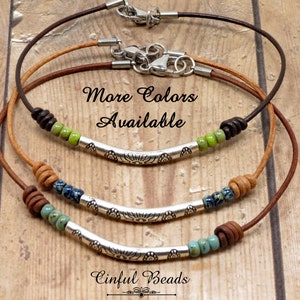 May include: Three leather cord necklaces with silver bar pendants and colorful beads. The necklaces are layered on a wooden surface. The text "More Colors Available" and "Cinfu Beads" are visible in the image.
