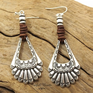 May include: A pair of silver dangle earrings with a brown leather cord and a silver bead at the top. The earrings have a fan-shaped design with intricate details.