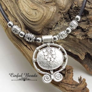 May include: A silver necklace with a round pendant featuring a hammered metal design and a Celtic knot motif. The necklace is made with black leather cord and silver beads.
