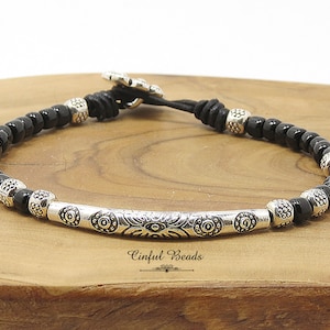 May include: Black beaded bracelet with silver accents and a decorative silver bar with a floral design. The bracelet has a sliding knot closure.