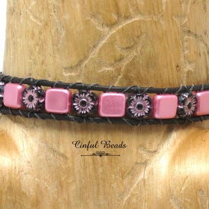 May include: A pink and black beaded bracelet with a leather strap. The beads are square with a floral design.
