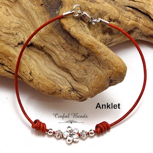 May include: Red leather ankle bracelet with a silver flower charm and silver beads. The bracelet is tied with a knot and has a silver clasp. "Cinfu Beads" is written on the bracelet.