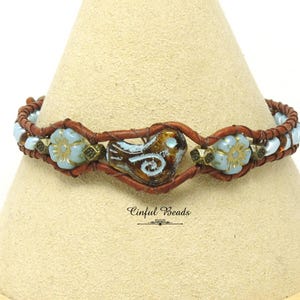 May include: A brown leather wrap bracelet featuring a central bird-shaped bead and light blue flower beads. The bracelet has a bohemian style, with a mix of textures and colors. The bracelet is displayed on a beige surface.