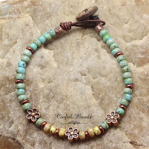 May include: A turquoise and yellow beaded bracelet with copper flower charms and a brown leather closure with a wooden button. The bracelet is on a brown, textured surface. "Cinfu Beads" is written on the surface.
