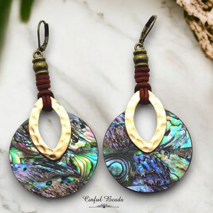 May include: A pair of dangle earrings featuring iridescent abalone shell discs with gold-tone hammered metal ovals. The earrings have a dark brown suede accent and a bronze-colored lever back closure. The earrings are designed with the text "Cinful Beads".