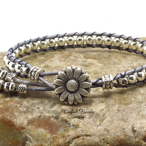 K&ouml;nnte beinhalten: Ein graues Lederarmband mit silbernen Perlen und einem silbernen G&auml;nsebl&uuml;mchen-Anh&auml;nger. Das Armband ist mit einer blauen Schnur gebunden.