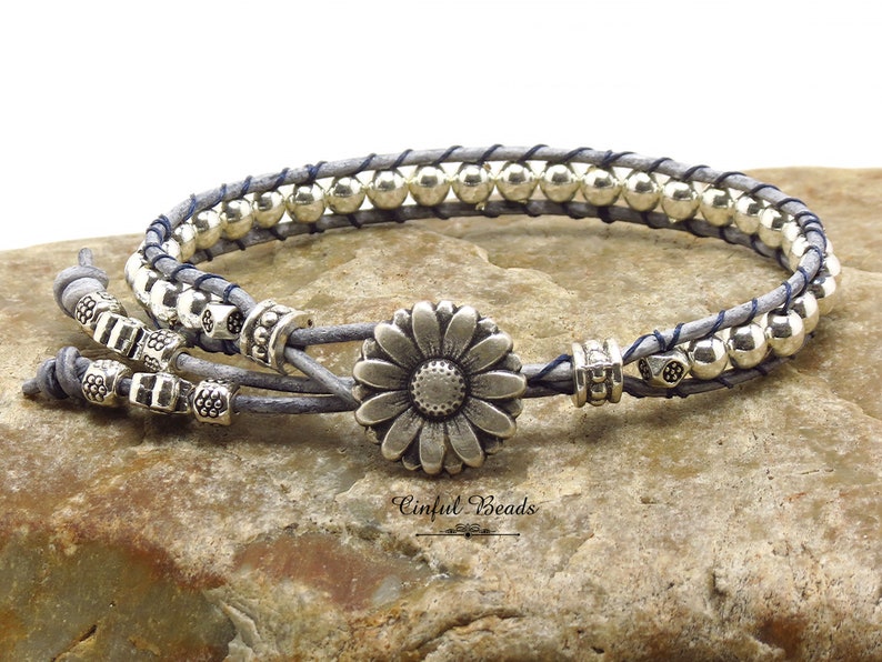K&ouml;nnte beinhalten: Ein graues Lederarmband mit silbernen Perlen und einem silbernen G&auml;nsebl&uuml;mchen-Anh&auml;nger. Das Armband ist mit einer blauen Schnur gebunden.