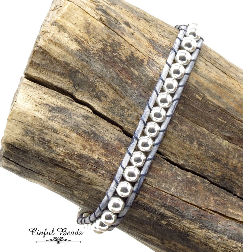 K&ouml;nnte beinhalten: Ein graues Lederarmband mit silbernen Perlen. Das Armband ist um ein St&uuml;ck Treibholz gewickelt. Der Text "Cinflul Beads" ist am unteren Rand des Bildes sichtbar.