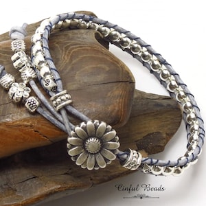 K&ouml;nnte beinhalten: Ein graues Leder-Wickelarmband mit silbernen Perlen und einem silbernen Blumenanh&auml;nger. Das Armband liegt auf einer Holzoberfl&auml;che. Der Text "Cinfu Beads" ist in der unteren rechten Ecke des Bildes sichtbar.