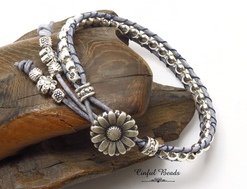K&ouml;nnte beinhalten: Ein graues Leder-Wickelarmband mit silbernen Perlen und einem silbernen Blumenanh&auml;nger. Das Armband liegt auf einer Holzoberfl&auml;che. Der Text "Cinfu Beads" ist in der unteren rechten Ecke des Bildes sichtbar.