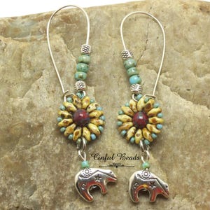 May include: A pair of dangle earrings featuring a sunflower design with a red center and yellow petals. The earrings have turquoise beads and silver bear charms. The earrings are displayed against a textured, tan background.