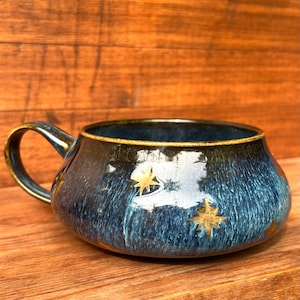 Könnte beinhalten: Ein breiter Keramikbecher mit dunkelblauer und schwarzer gesprenkelter Glasur und goldenen Sternakzenten. Der Becher hat einen niedrigen, abgerundeten Körper, eine weite Öffnung und einen einzelnen geschwungenen Henkel auf der linken Seite.