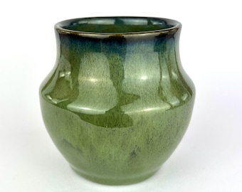 May include: A handcrafted ceramic vase with a glossy green glaze. The vase has a rounded body and a slightly flared rim, with a darker green-blue hue at the top. The surface has subtle variations in color and texture.