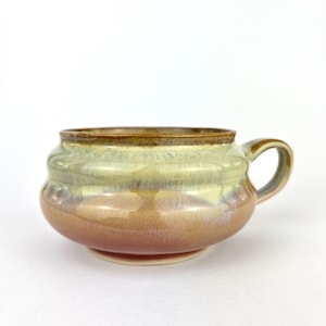May include: A handmade ceramic soup bowl with a handle. The bowl features a rounded shape with a footed base and a brown rim. The exterior has a gradient of colors, including peach, tan, and green.