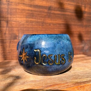 Könnte beinhalten: Eine blaue Keramiktasse mit Henkel, mit dem goldenen Text "Jesus is" und einem goldenen Stern-Design. Die Tasse hat eine glänzende Oberfläche und steht vor einem Holzgrund.