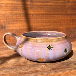 May include: A wide, low-profile ceramic mug with a rounded body and a single handle. The exterior features a glossy, blended glaze of soft lavender, pink, and cream tones, decorated with several small, metallic gold star-shaped accents.