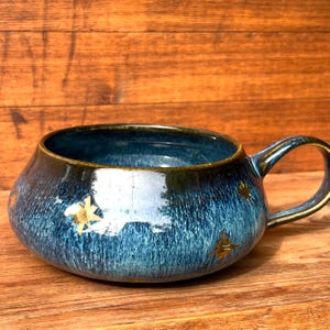 May include: A wide, low-profile ceramic mug with a glossy, deep blue glaze featuring a vertical streaked texture. The exterior is decorated with small, metallic gold star-shaped accents scattered across the surface, and it has a single handle.