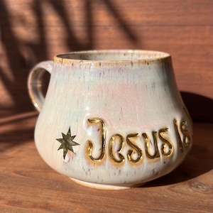 Könnte beinhalten: Cremefarbene Keramiktasse mit runder Form und Henkel. Die Tasse zeigt den goldfarbenen Text "Jesus Is" und ein Stern-Design. Der Rand und der Henkel haben einen goldfarbenen Besatz.