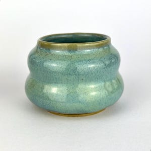 Könnte beinhalten: Eine kleine, handgefertigte Keramikschale mit einer einzigartigen, abgerundeten Form. Die Schale hat eine gesprenkelte, türkisfarbene Glasur mit einem Hauch von Grün am Rand und am Boden. Die Keramik ist wahrscheinlich handgefertigt, was ihren handwerklichen Reiz verstärkt.
