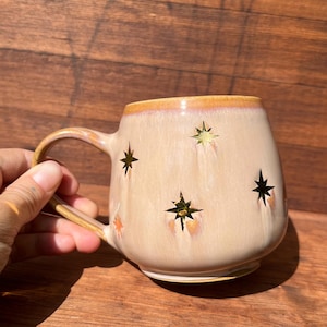 Op de afbeelding: Een beige keramische mok met een afgeronde vorm en een grote handgreep. De mok heeft stervormige uitsparingen en een goudkleurige rand. De mok staat op een houten oppervlak.