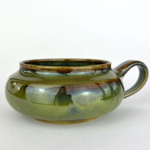 May include: A handmade ceramic bowl with a handle, featuring a glossy, iridescent green glaze. The bowl has a rounded shape with a brown rim and handle, perfect for serving soups or stews.