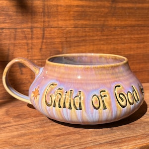 Może przedstawiać: Szeroki, niski kubek ceramiczny z błyszczącym, opalizującym szkliwem w odcieniach lawendy, różu i złota. Napis "Child of God" jest wytłoczony złotymi literami z przodu, otoczony po obu stronach małymi ikonami złotych gwiazdek.