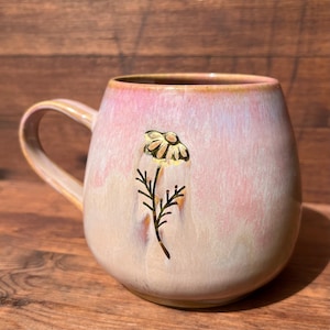 May include: A ceramic mug with a rounded shape and a handle. The mug has a pink and cream glaze with a gold-colored flower and stem design. The mug is sitting on a wooden surface.