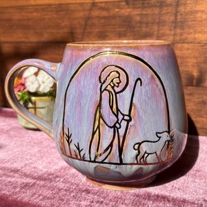 Könnte beinhalten: Ein Keramikbecher mit einer glänzenden, lavendel- und hellblauen Glasur. Die Vorderseite zeigt eine dunkle Umrisszeichnung einer Figur mit Heiligenschein, die einen Stab hält, begleitet von einem kleinen Lamm, eingerahmt von einem dunklen Bogen.