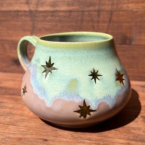 May include: A ceramic mug with a rounded shape and a handle. The mug features a light green and tan color scheme with star cutouts. The interior is a light green color. The mug is sitting on a wooden surface.