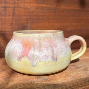 May include: A handmade ceramic mug with a rounded body and a curved handle. The mug features a blend of colors, including yellow, peach, and white, creating a unique, marbled effect. The mug is suitable for coffee, tea, or other beverages.
