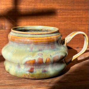 Könnte beinhalten: Ein breiter, flacher Keramikbecher mit einem abgerundeten, bauchigen Körper und einer markanten horizontalen Wulst. Die Glasur zeigt eine Mischung aus Salbeigrün, warmem Bernstein und tiefen Brauntönen mit glänzender Oberfläche und einem Henkel.