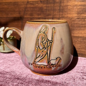Może przedstawiać: Ceramiczny kubek o zaokrąglonym kształcie z błyszczącym, wielokolorowym szkliwem w odcieniach beżu, różu i brązu. Z przodu znajduje się wypukły, ciemno obrysowany relief przedstawiający postać w szatach trzymającą laskę pasterską oraz małą owcę.