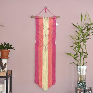 Puede incluir: Un tapiz de pared de macramé hecho a mano en tonos rosa, melocotón y crema. La decoración de la pared presenta un patrón de diamantes geométricos y flecos largos. Está suspendido de un pasador de madera natural y un cordón rosa.