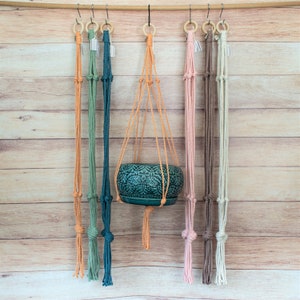 May include: A collection of macrame plant hangers in various colors, including orange, green, teal, pink, and brown. One hanger holds a teal ceramic pot with a detailed pattern. The hangers are suspended from a wooden rod with metal hooks.