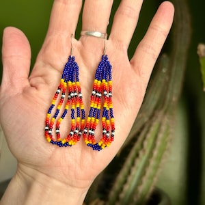 Puede incluir: Pendientes de cuentas con diseño de lágrima. Los pendientes presentan una combinación de cuentas azules, amarillas, rojas, negras y blancas. La parte superior es principalmente azul, con los otros colores debajo. Los pendientes cuelgan de ganchos plateados.