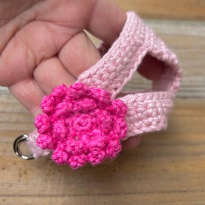 May include: A light pink crocheted harness with a bright pink flower accent. The harness has a silver metal ring attached. The flower is a vibrant fuchsia color, and the harness is held in a hand.