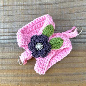 May include: A pink crocheted headband with a dark purple flower and green leaves. The flower has a rhinestone center. The headband has a metal ring and a pink plastic clip attached.