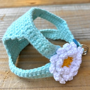 May include: A light blue crochet pet harness with a white daisy flower accent. The harness is made of soft yarn and features a metal ring for leash attachment. The daisy has a yellow center.