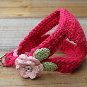 May include: A pink crocheted dog harness with a pink flower and green leaves. The harness has a metal D-ring for attaching a leash.