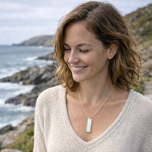 May include: A silver pendant necklace with a textured rectangular pendant worn by a woman. The necklace hangs from a fine chain. She wears a cream-coloured sweater, with a coastal backdrop of the sea and rocks.