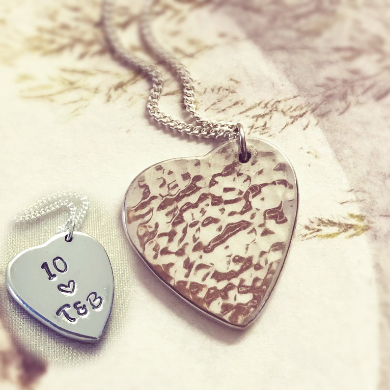 May include: Two silver heart-shaped pendants on a silver chain. The larger pendant has a hammered texture. The smaller pendant has the inscription "10 &hearts; T&B".