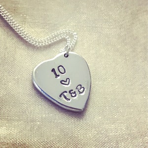 May include: Silver heart-shaped pendant necklace with a chain. The pendant has the text "10" with a heart symbol and "T&B" engraved on it.